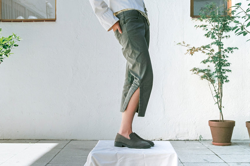 Lifestyle photo of a person wearing Legacy Slipper Suede Grey Walnut loafers with a dark green midi skirt, standing near a potted plant