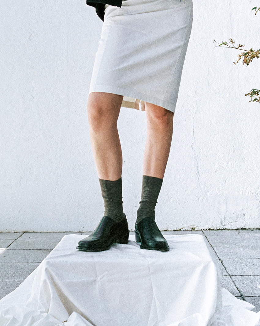 Lifestyle close-up of legs and feet wearing Pedal Loafer Pull-up Leather Black shoes with dark green socks and a grey skirt
